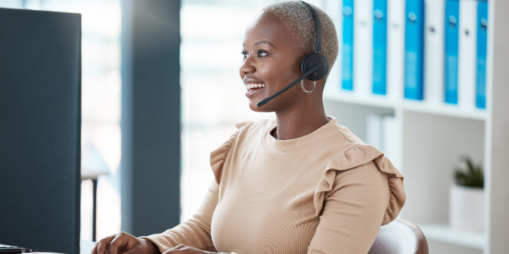 A smiling woman wears a headset while seated at a computer in an office.